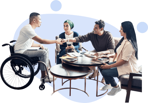 Four colleagues collaborating at a table, including a man using a wheelchair, exchanging a handshake.