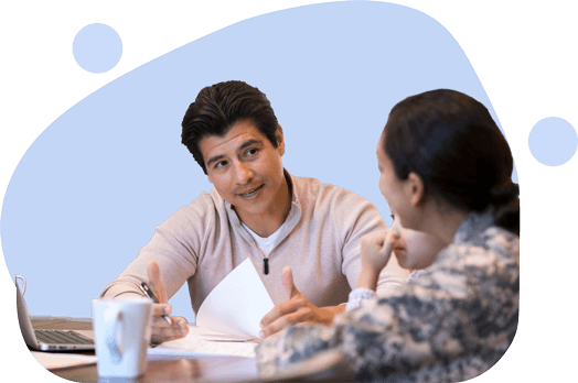 Two colleagues in discussion at a table, one explaining while the other listens.